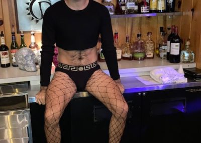 Person wearing all black risqué costume sits on top of bar counter Halloween 2021 Party.