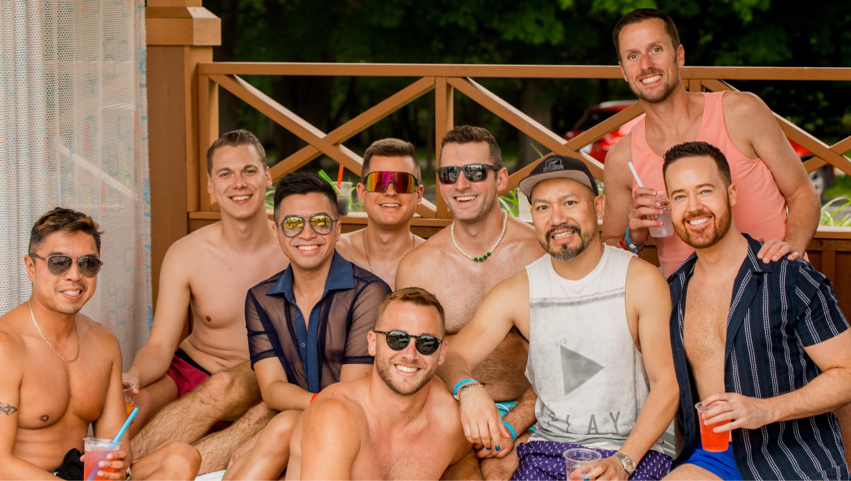 Eight people in sunglasses and swimsuits pose in a cabana.