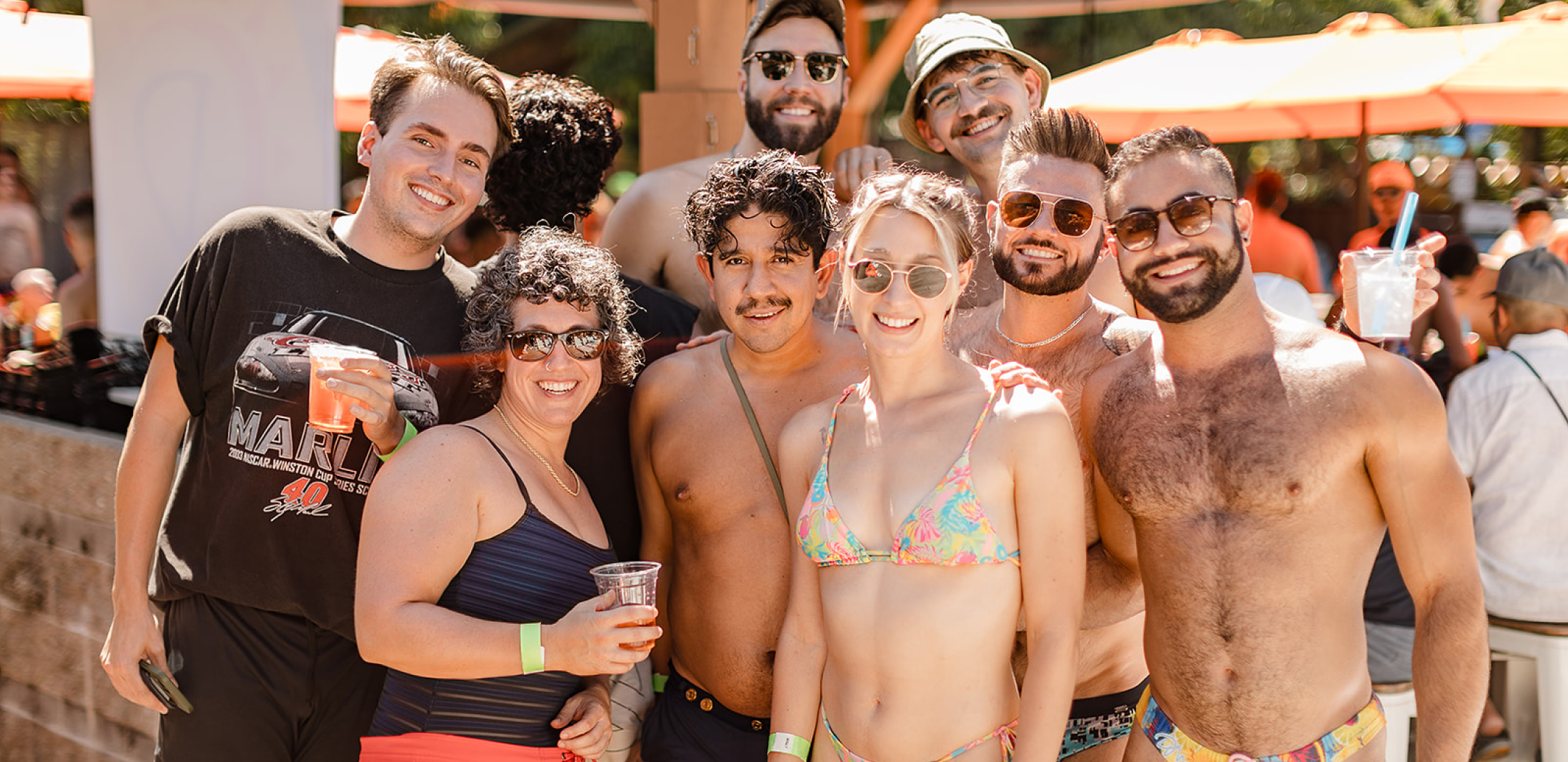 A group of smiling people in swimsuits enjoying a sunny day at an outdoor venue. They are holding drinks, wearing sunglasses, and appear relaxed and happy.