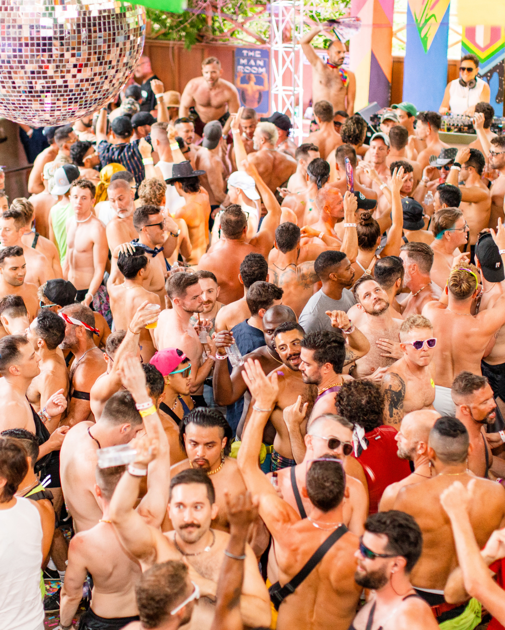 A vibrant crowd of shirtless people at a Tea Dance on the Deck, with everyone dancing energetically under a large disco ball.
