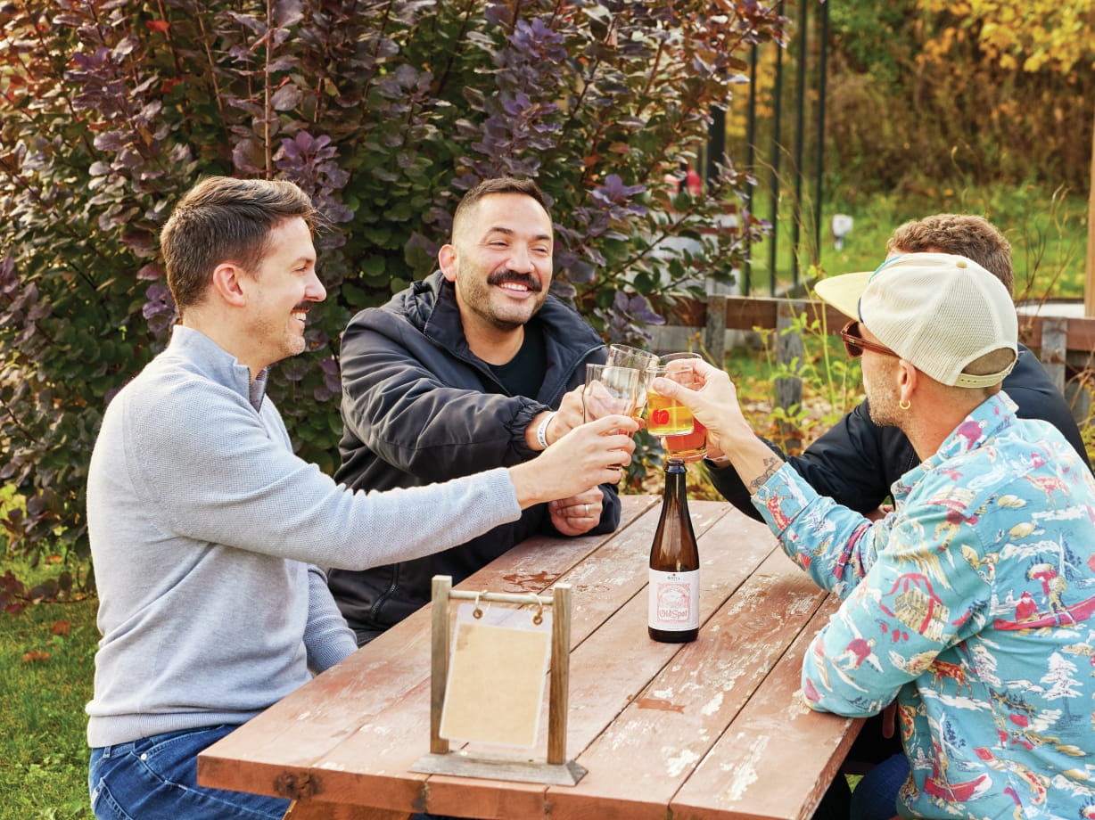 Friends toast at an outdoor table Virtue Cider, enjoying drinks and laughter in a lush outdoor setting.