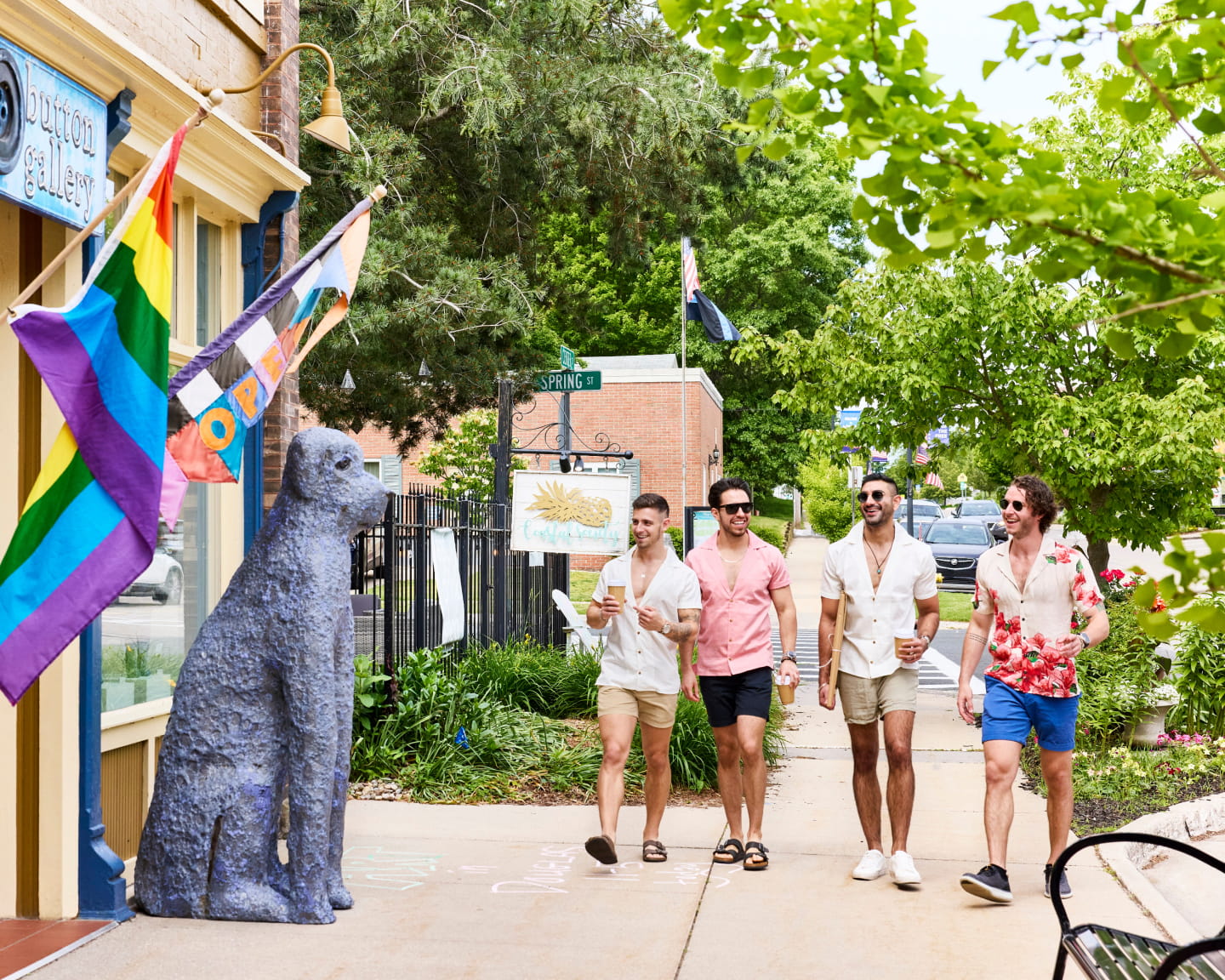 Four men in summer attire walk past rainbow flags and a dog statue on a sunny street, holding drinks and smiling.