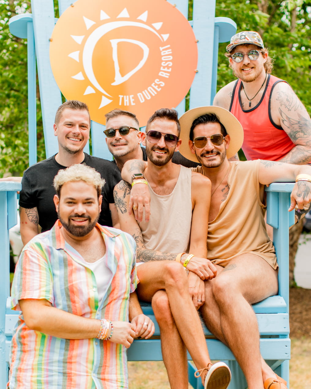 Six friends sit on a large blue chair at The Dunes Resort, smiling and enjoying a sunny day outdoors.