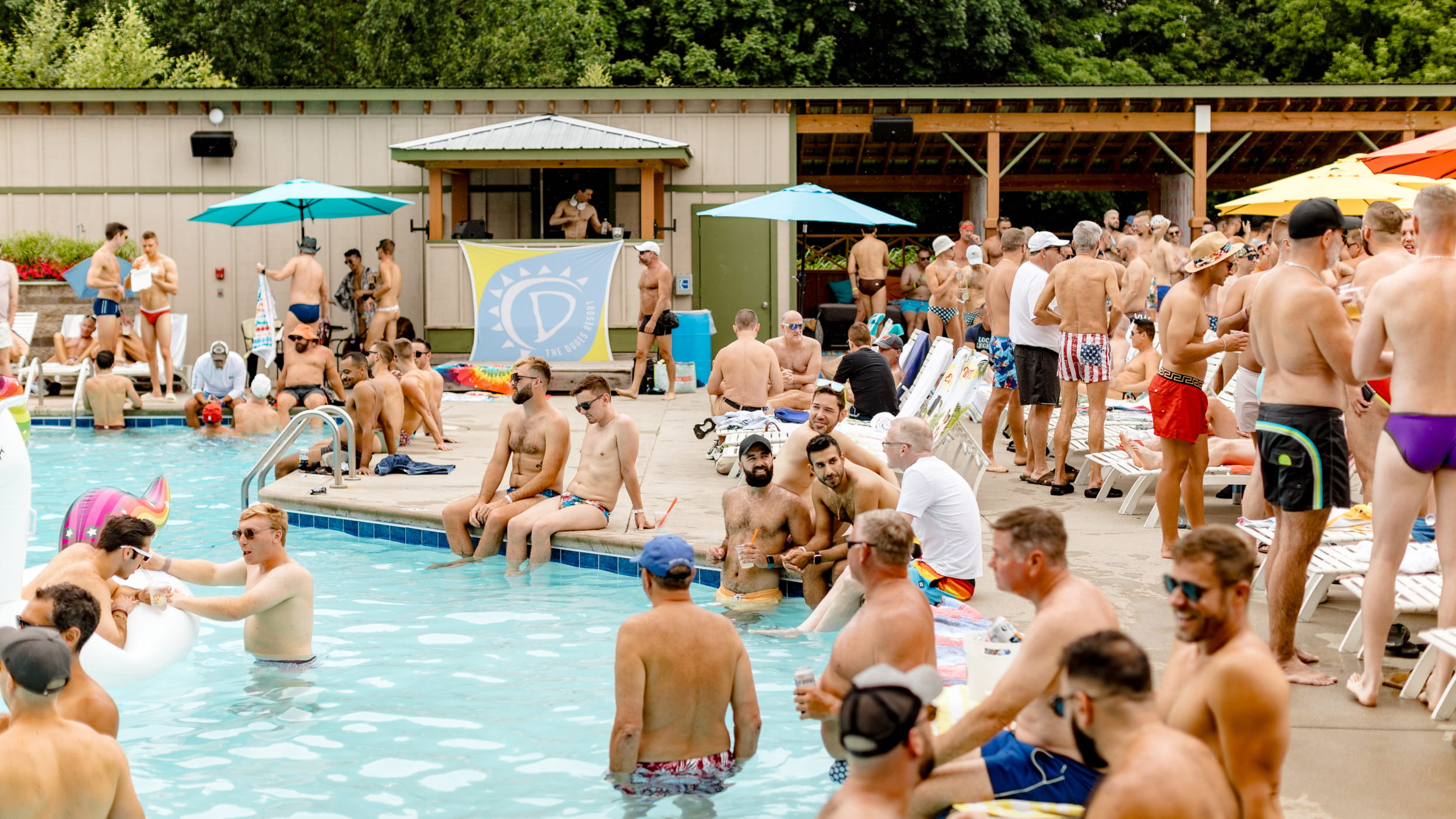 People enjoy a lively pool party at the Pool + Splash Bar, with colorful umbrellas and a vibrant atmosphere.
